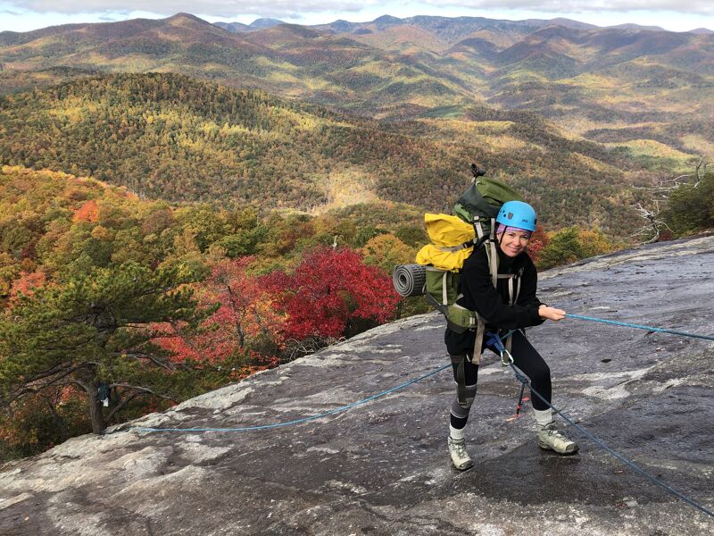 A woman is rappelling down a rock face with a large backpack. She is wearing a helmet and safety gear. The background shows a valley with trees in fall colors. The sky is cloudy.
